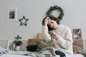 A woman sits in her bed wearing an oversize sweater looking at her phone concerned