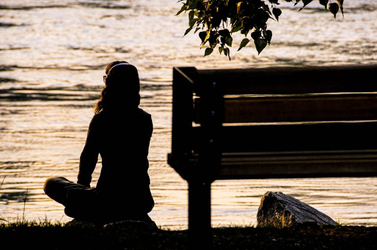 A woman with her back to the camera, sitting beside a bench cross legged facing the lake