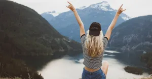 woman sitting on a rock facing the lake and mountains with her black hat on backwards and arms reaching up in triumph